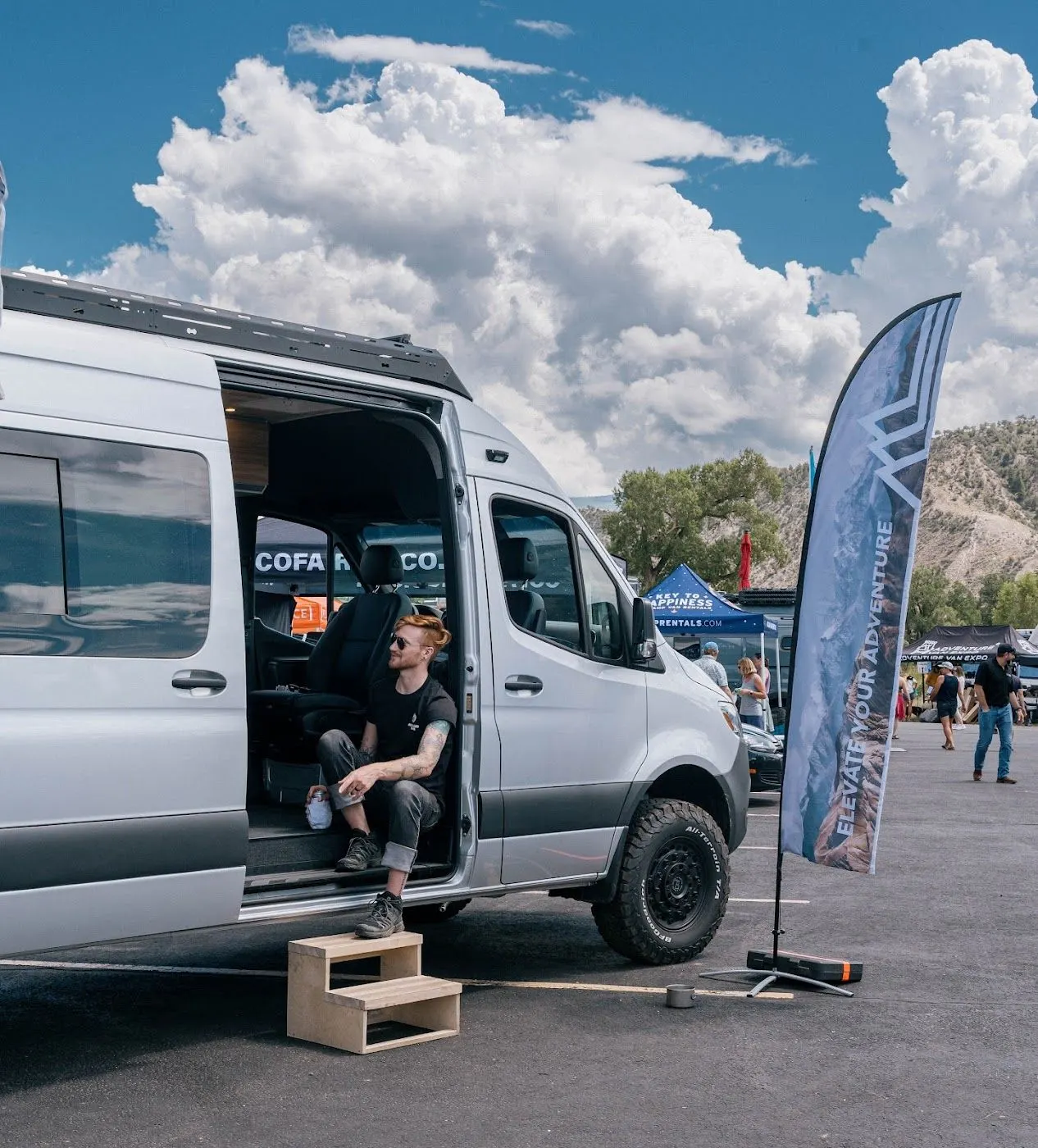 An owner sitting in the open side door of his Highland Vans Sprinter at a mountain show, beside a Highland Vans flag reading Elevate Your Adventure