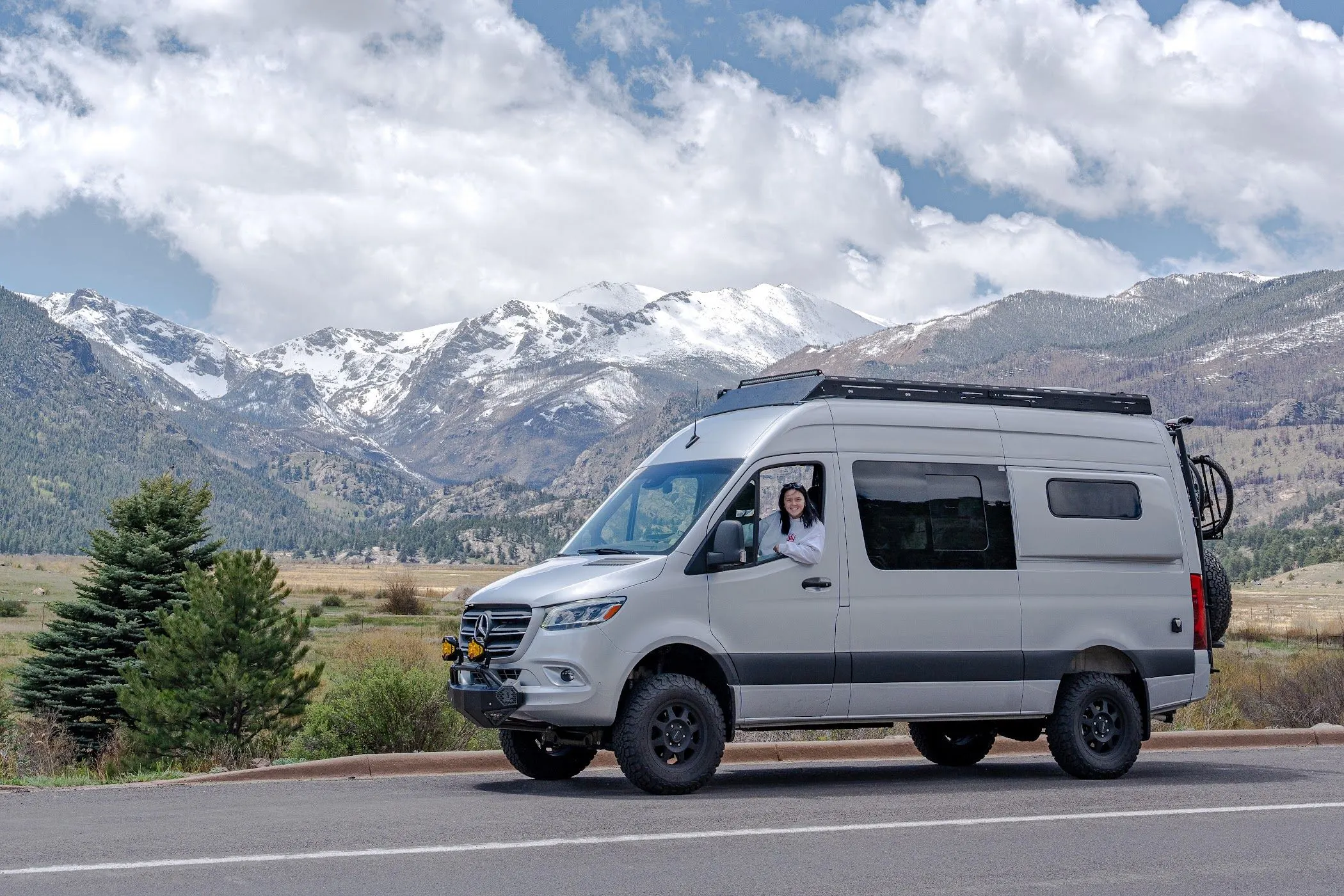 A silver Highland Vans Sprinter parked on a mountain road below snow-capped peaks, with a person leaning out of the open driver's window