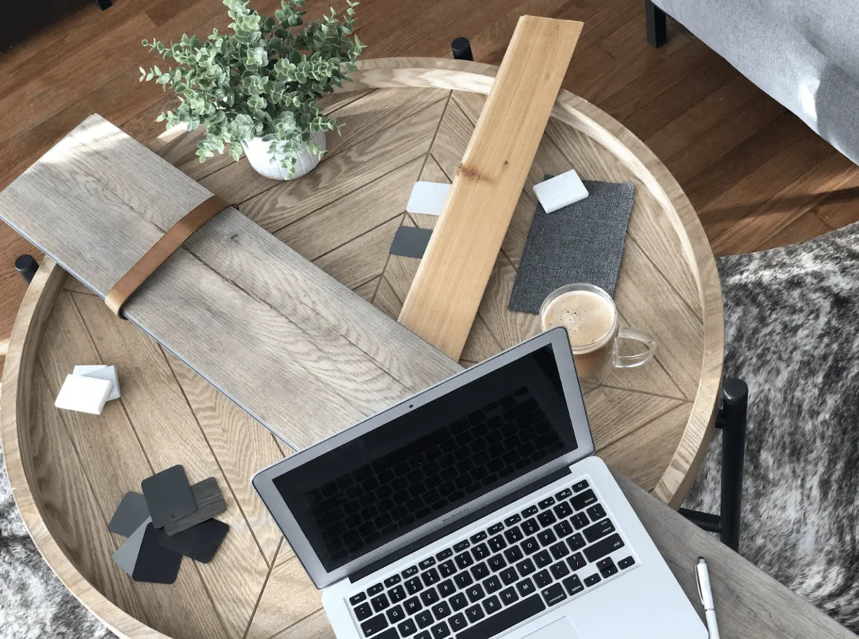Wood veneer planks, fabric swatches and grey and white colour chips laid out on a round oak table beside an open laptop and a coffee