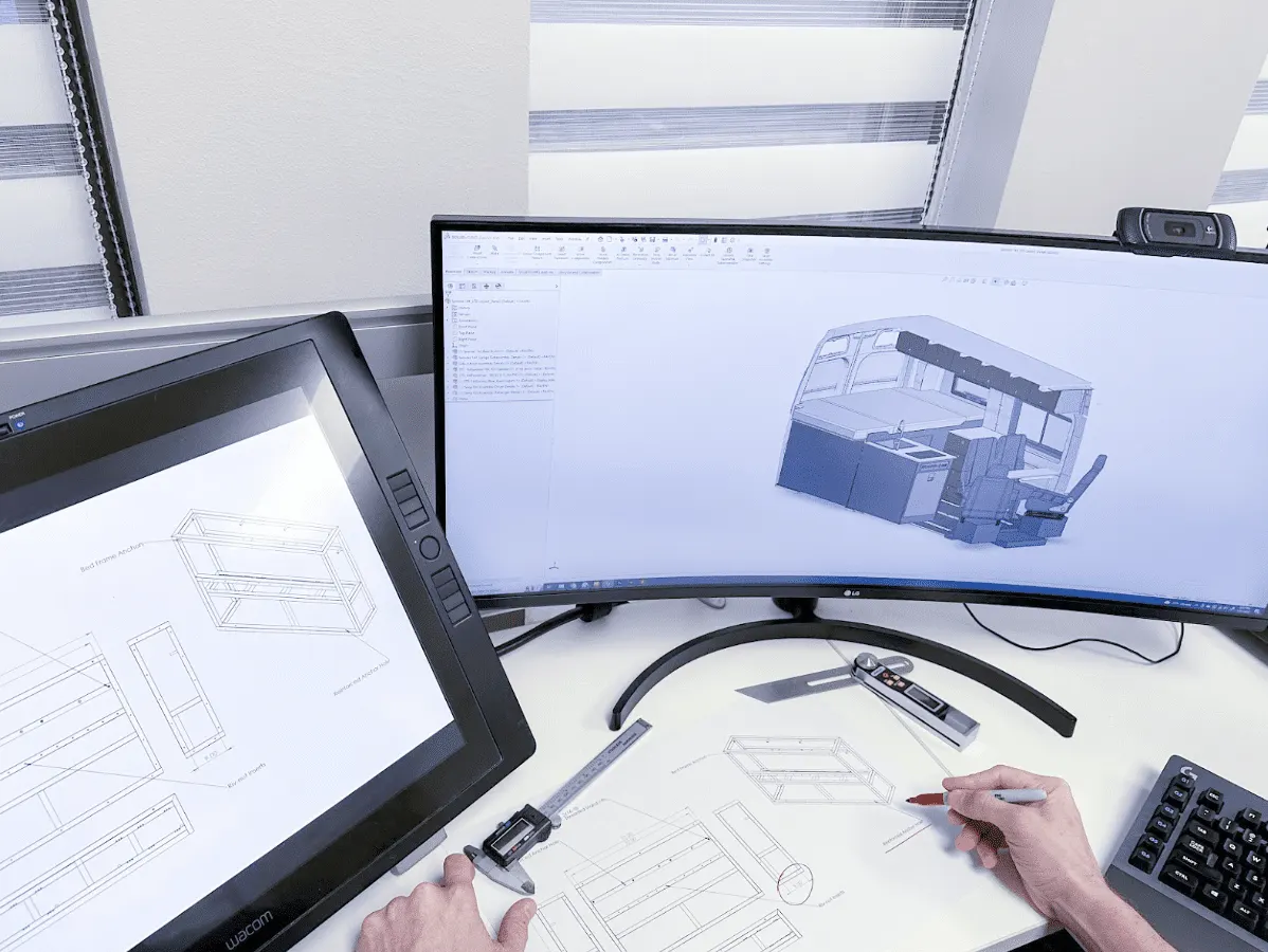 A designer’s desk with a van interior modelled in CAD on a curved monitor, a bed-frame drawing on a pen display, and two hands working over printed drawings with digital calipers and a marker