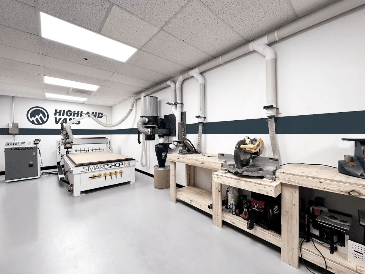 The Highland Vans CNC room: a flatbed router and its dust extraction beside timber benches and a mitre saw, with the Highland Vans logo painted on the white wall