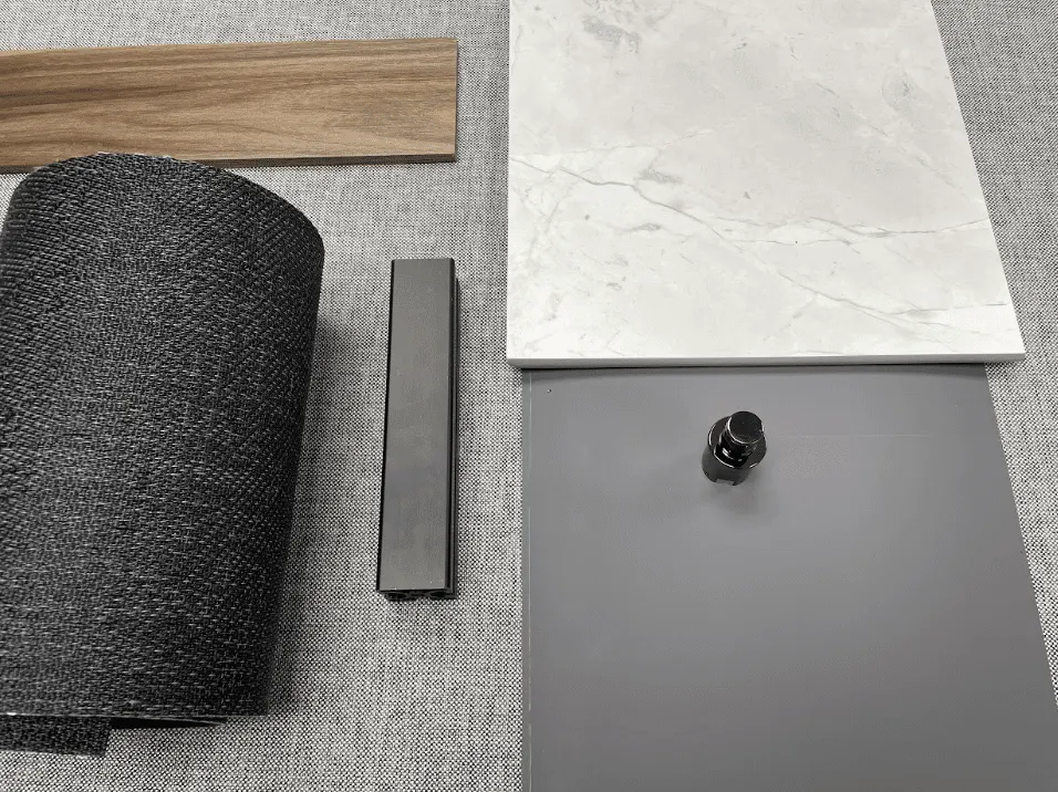 Material samples on a grey linen ground: a wood-grain plank, a roll of dark woven upholstery fabric, a brushed aluminium extrusion, a white marble-pattern solid surface and a grey cabinet door with a chrome knob