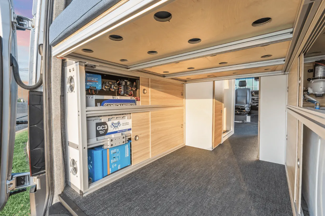The rear of a finished conversion with a cabinet panel removed, showing the electrical bay: blue Victron charger and inverter units, a lithium battery, fused distribution blocks and cooling fans, under a birch ply ceiling with recessed lights