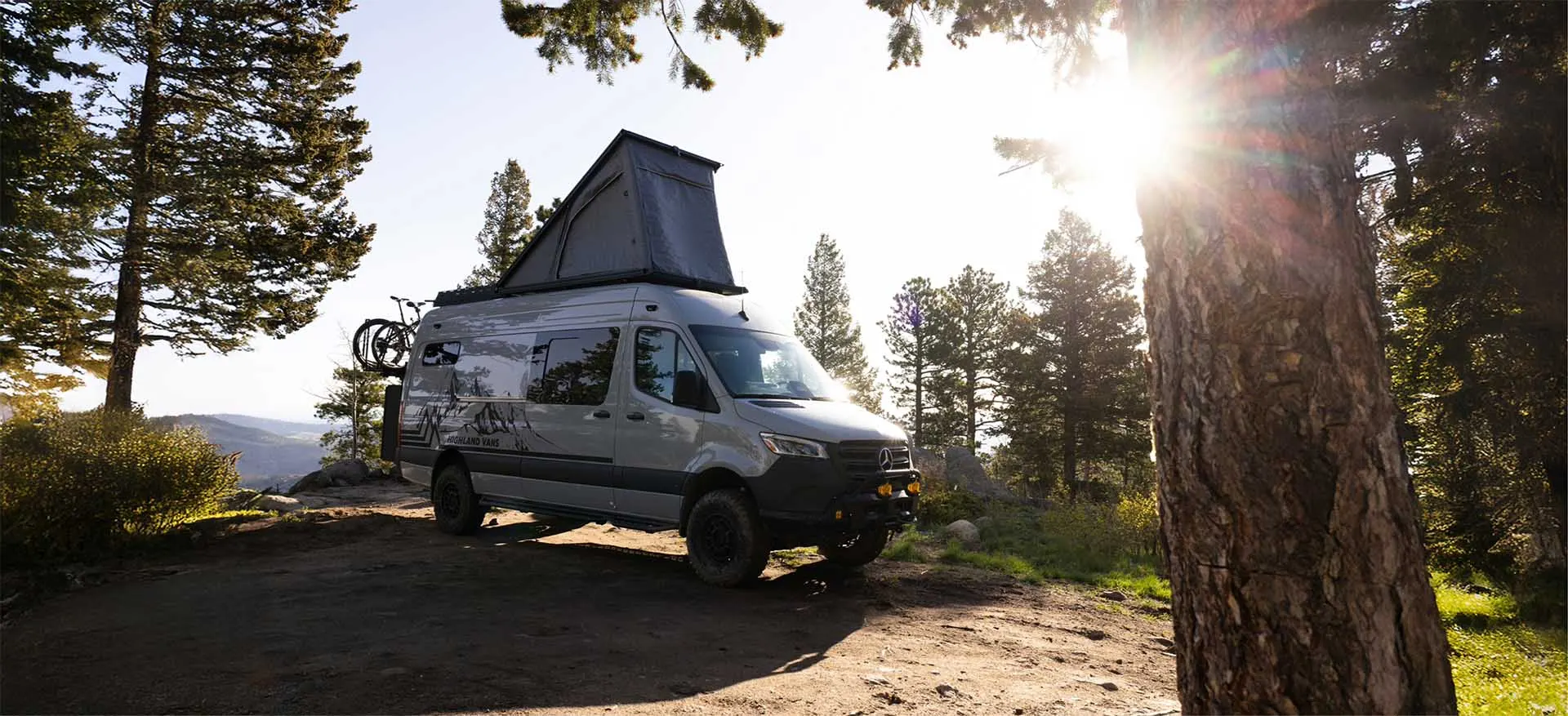 A Highland Vans Sprinter with its pop-top roof raised, parked at a forest pull-off with the sun breaking through the pines behind it