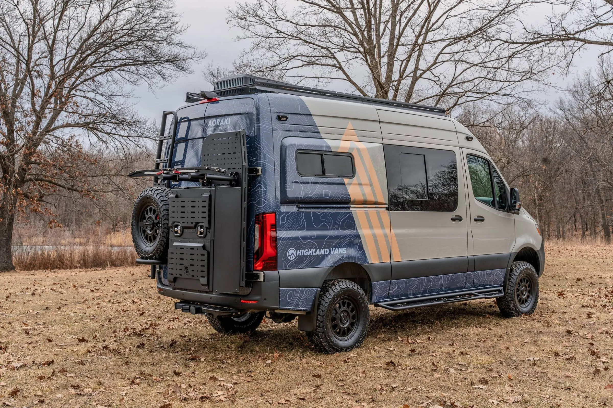 A Highland Vans Sprinter in blue and grey topographic livery parked in winter woodland, its rear ladder, gear carrier and spare wheel visible on the back doors