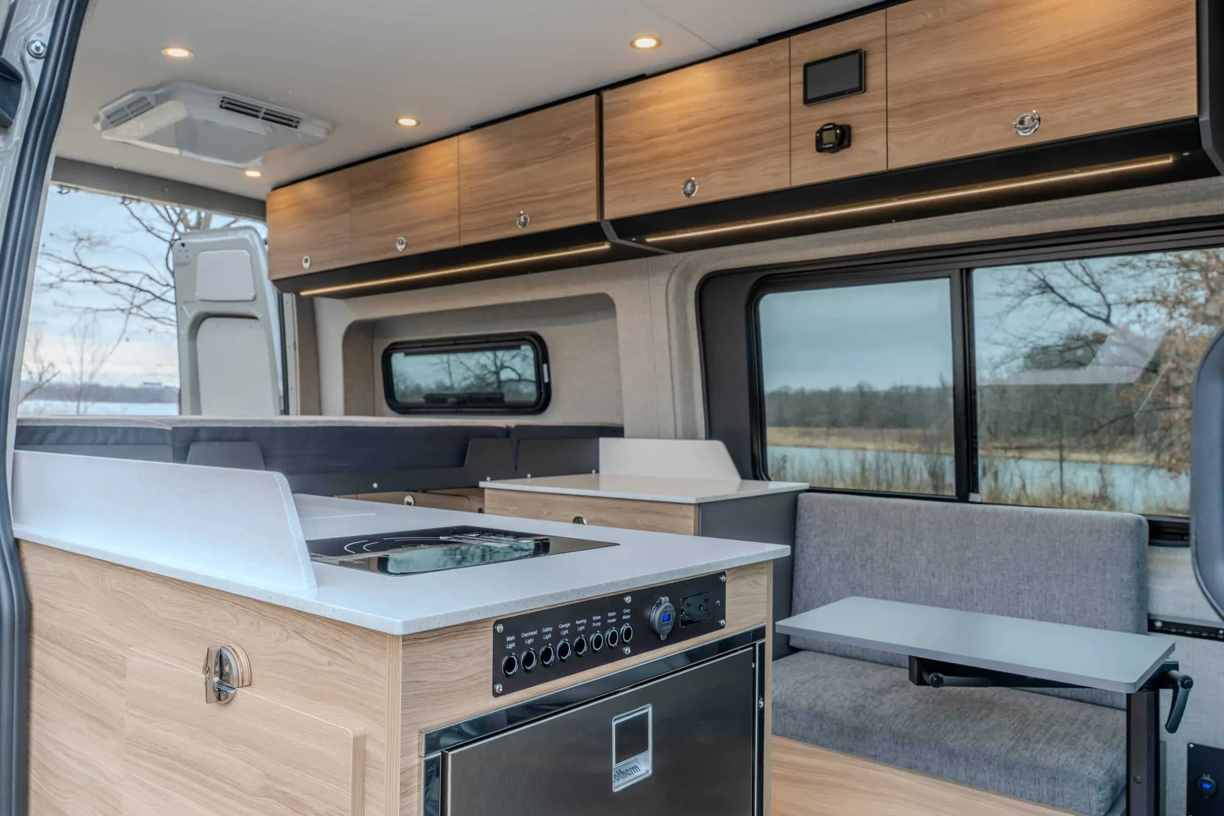 The galley of a finished Highland Vans conversion: oak-faced cabinetry, a pale stone counter with a flush induction hob, a labelled switch panel for the lights, water pump and water heater, and a bench seat at a window onto open water