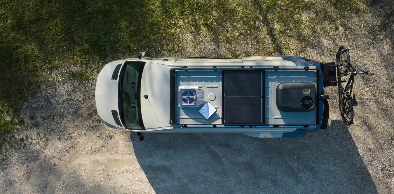 A Highland Vans Sprinter photographed from directly above on a gravel track, its roof carrying a rack, a solar panel, a roof fan, an air conditioner and a bicycle on the rear carrier