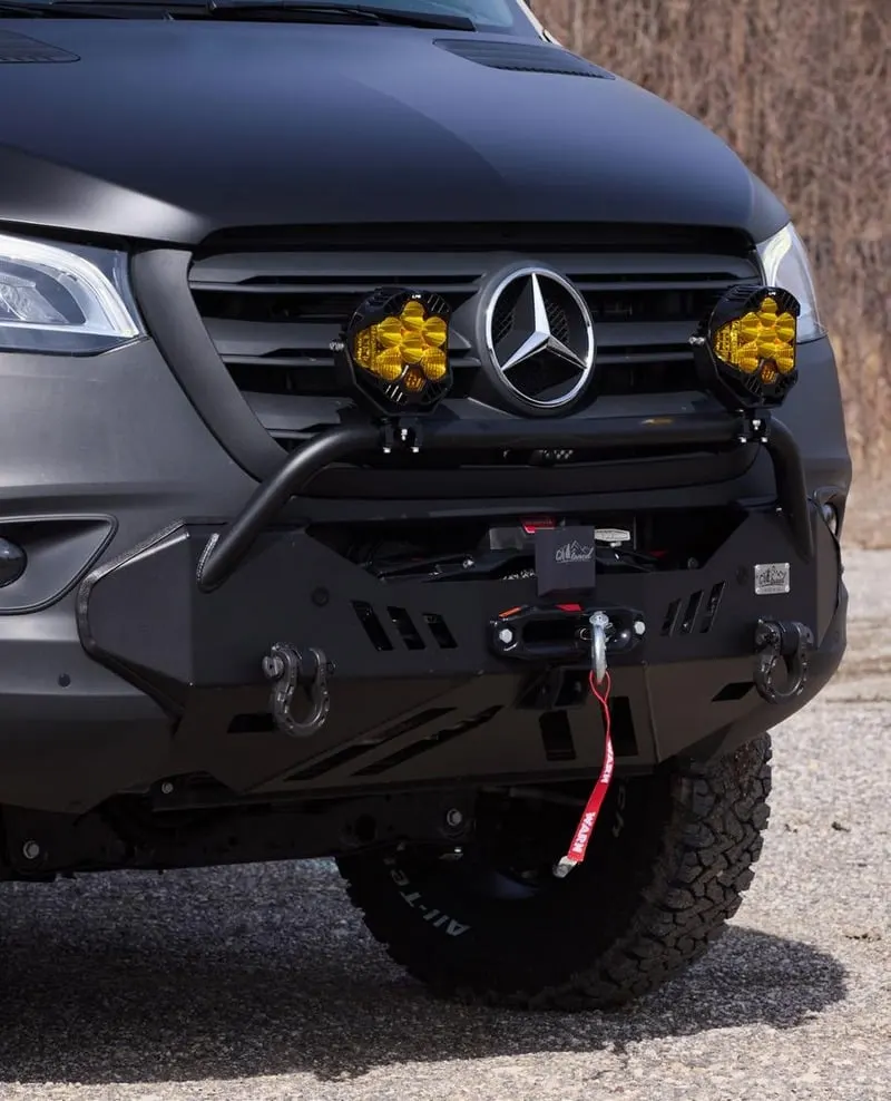 The front of a matte black Sprinter with an off-road bumper, a recessed winch and two yellow-lensed auxiliary lights beside the grille