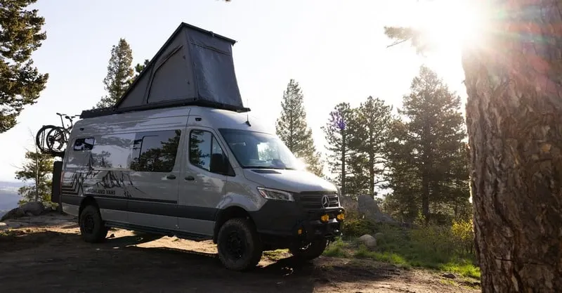 A Highland Vans Sprinter with its pop-top roof raised, parked among pines on a ridge with the sun low behind the trees