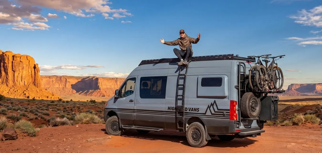 A Highland Vans Sprinter parked on red sand below the buttes of Monument Valley, a person sitting on its roof rack with both arms raised, two mountain bikes on the rear carrier