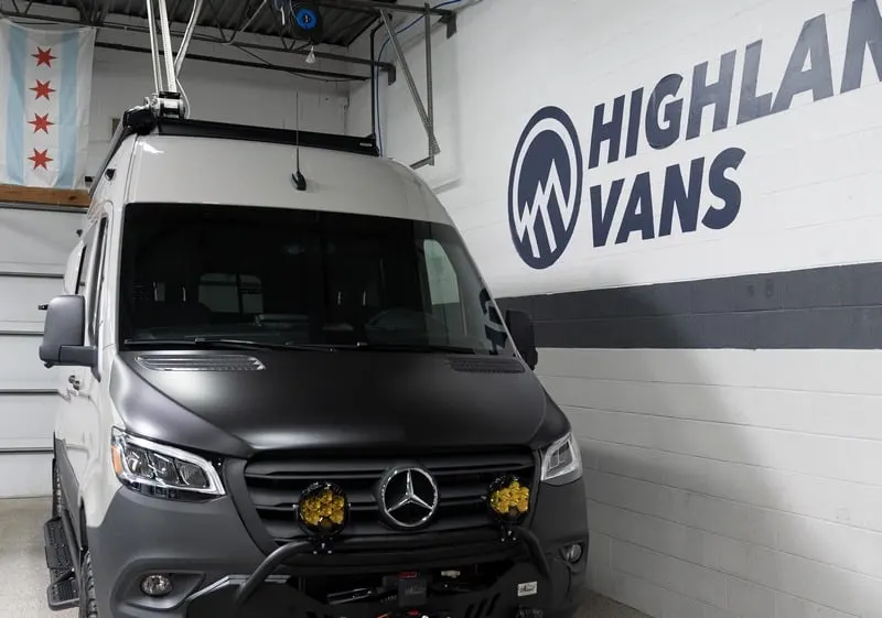 The front of a Highland Vans Sprinter with a matte black bonnet, a bull bar and two auxiliary lights, parked inside the shop beside the Highland Vans wordmark painted across the wall