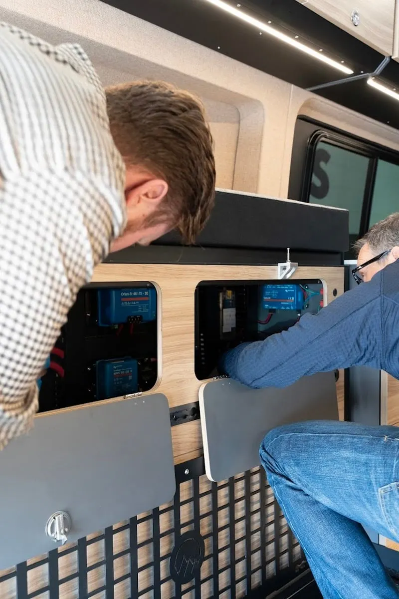 Two people at the open side door of a van, working on its electrical bay: a Victron converter and charge controller behind one opened panel and the distribution board behind another