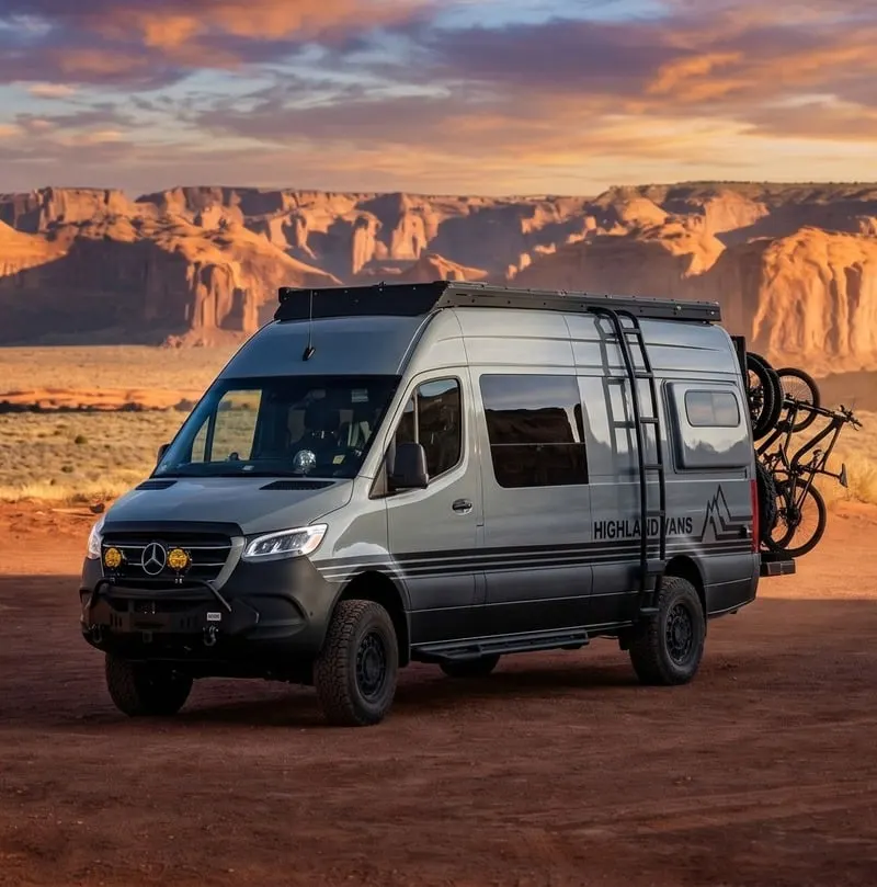 The front three-quarter view of a Highland Vans Sprinter on red dirt at sunset, sandstone mesas behind it and auxiliary lights in the bumper