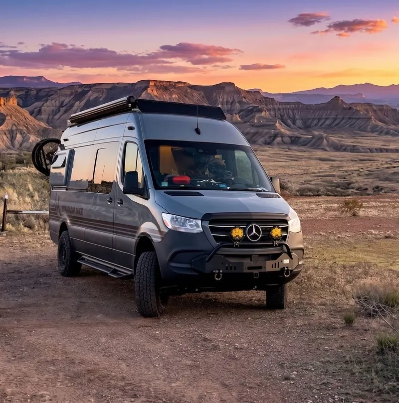 The front three-quarter view of a Highland Vans Sprinter at dusk on a high desert pull-off, layered badlands and an orange sky behind it