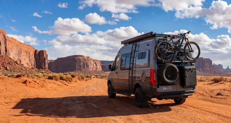 The rear three-quarter view of a Highland Vans Sprinter on a red dirt road running away between the buttes of Monument Valley