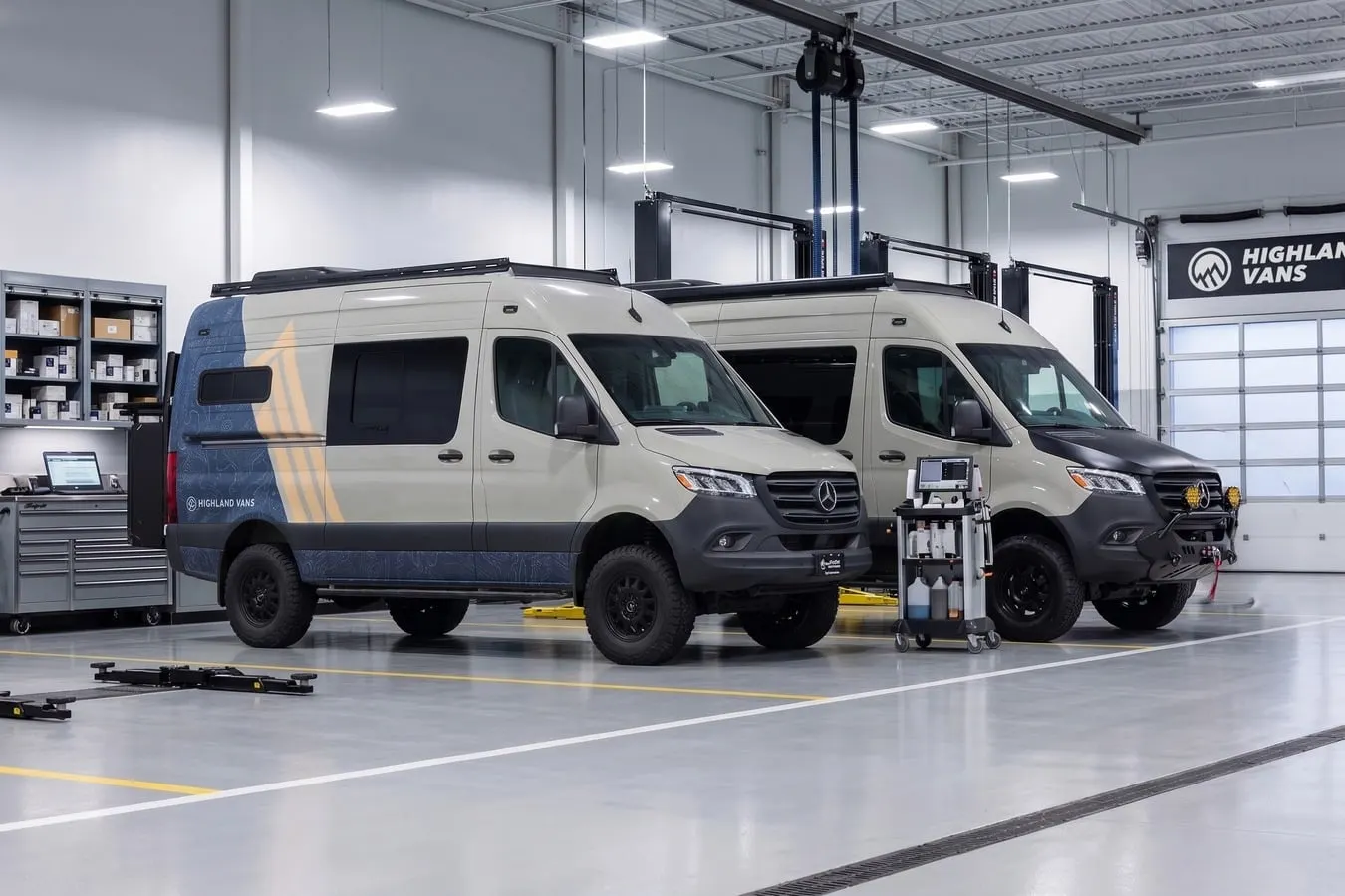 Two Highland Vans Sprinters inside the Elk Grove Village shop, one standing on a lift beside a mobile service cart, with a parts rack and a workbench along the left wall and the Highland Vans wordmark above the roller door