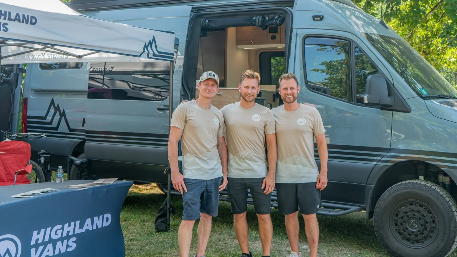 Three of the Highland Vans team standing shoulder to shoulder in front of an open Sprinter conversion at an outdoor show, under a Highland Vans canopy