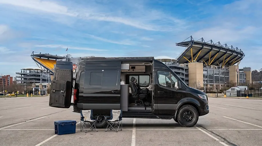 The same Kinabalu parked outside a stadium, sliding door open on the belted rear seats, two camp chairs and a cooler set out beside it