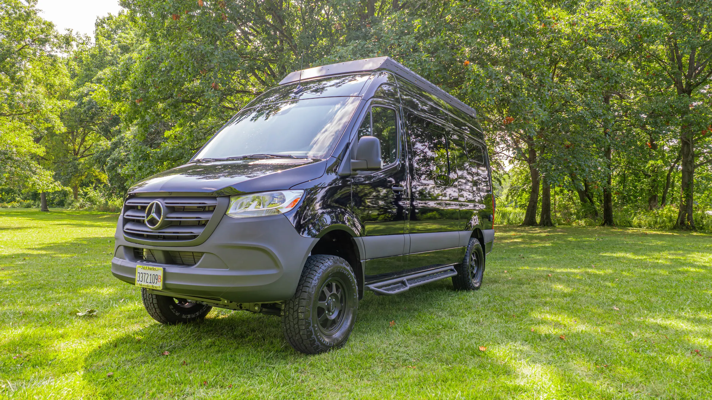 Front three-quarter view of the black Kinabalu on grass, with the low roof rack, side steps and all-terrain tyres on black wheels