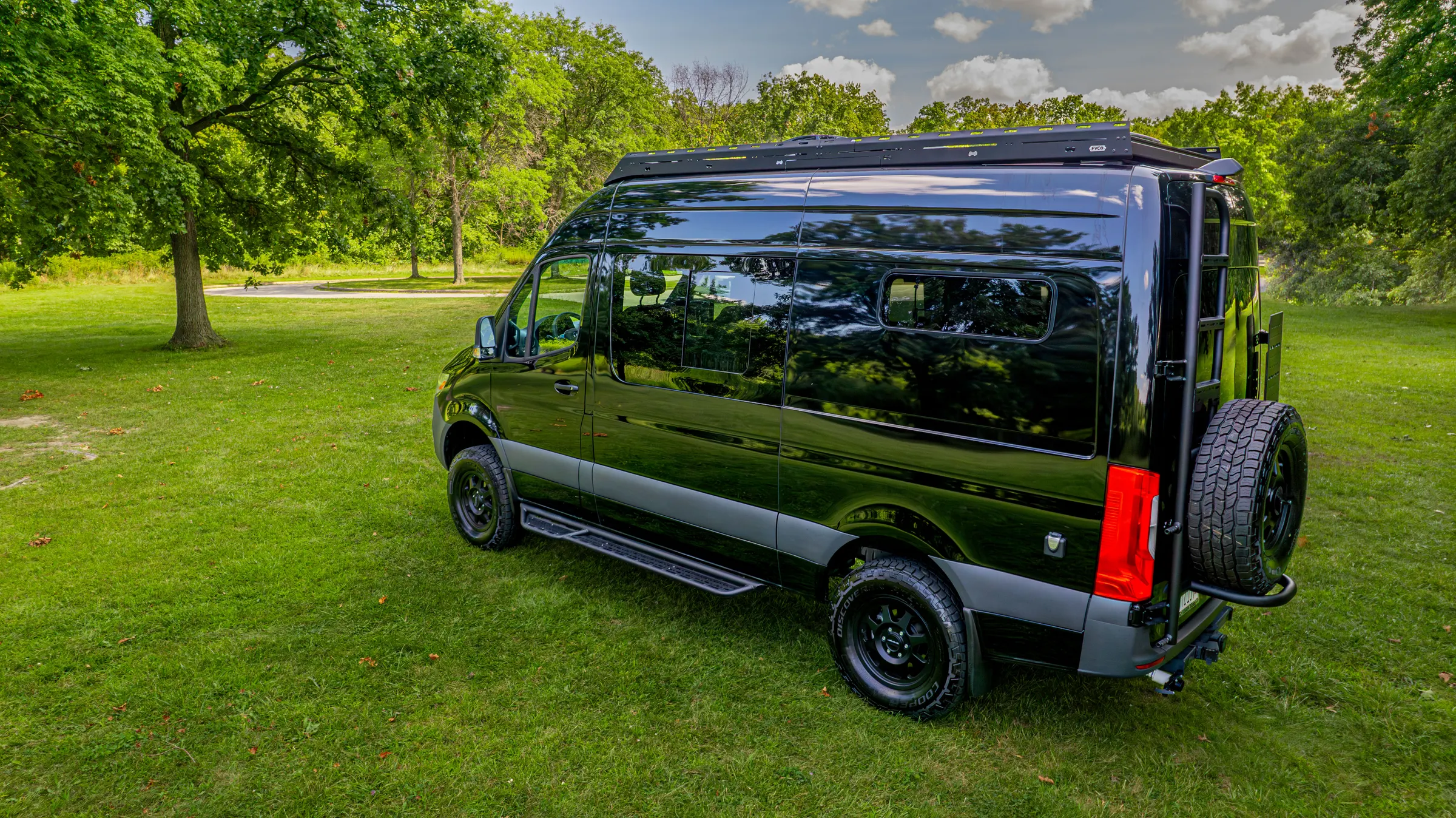 Rear three-quarter view on grass, showing the roof rack, the rear ladder and the spare wheel on its swing-out carrier
