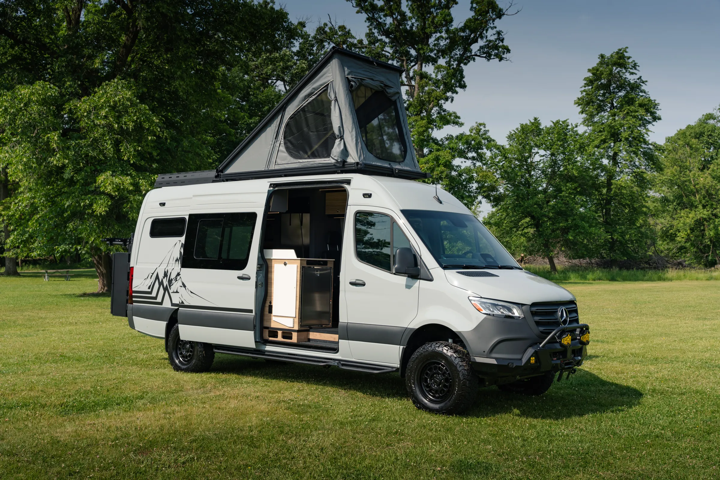 The pop-top roof tent raised and the sliding door open, seen from the front three-quarter with the bull bar and its auxiliary lights ahead