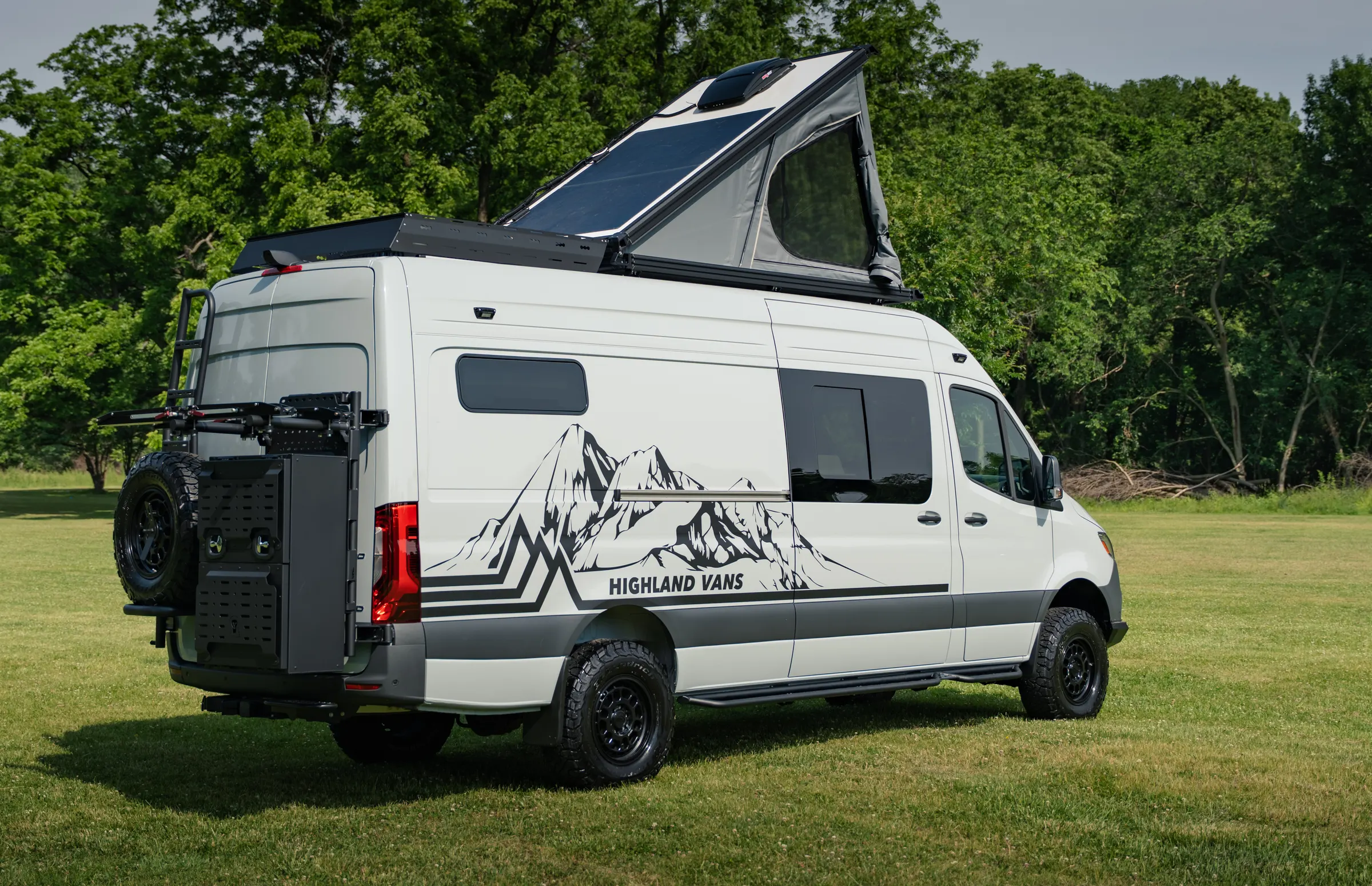 Rear three-quarter with the roof tent raised, showing the swing-out spare wheel, the perforated cargo panel beside it and the rear ladder