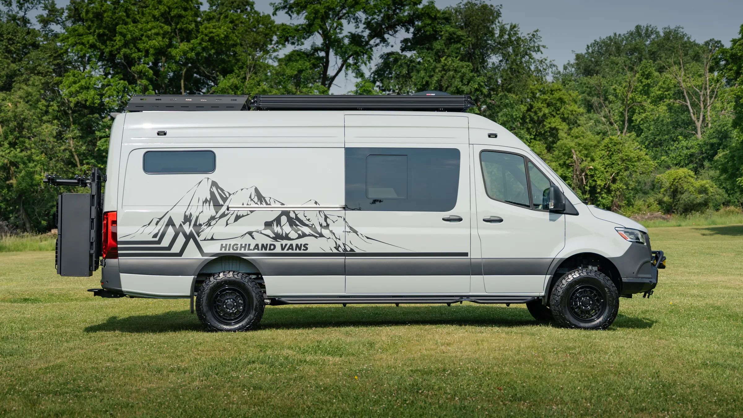 Full side profile on grass: the long-wheelbase body in white over a grey band carrying the Highland Vans mountain graphic, with a low roof rack, a side step and all-terrain tyres on black wheels