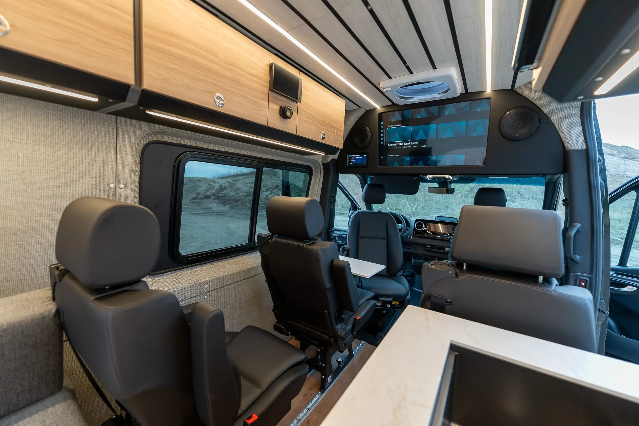 The cabin of a finished Highland Vans Sprinter looking forward from the rear: a slatted wood ceiling and upper cabinets, woven fabric wall panels, leather swivel seats and a stone counter