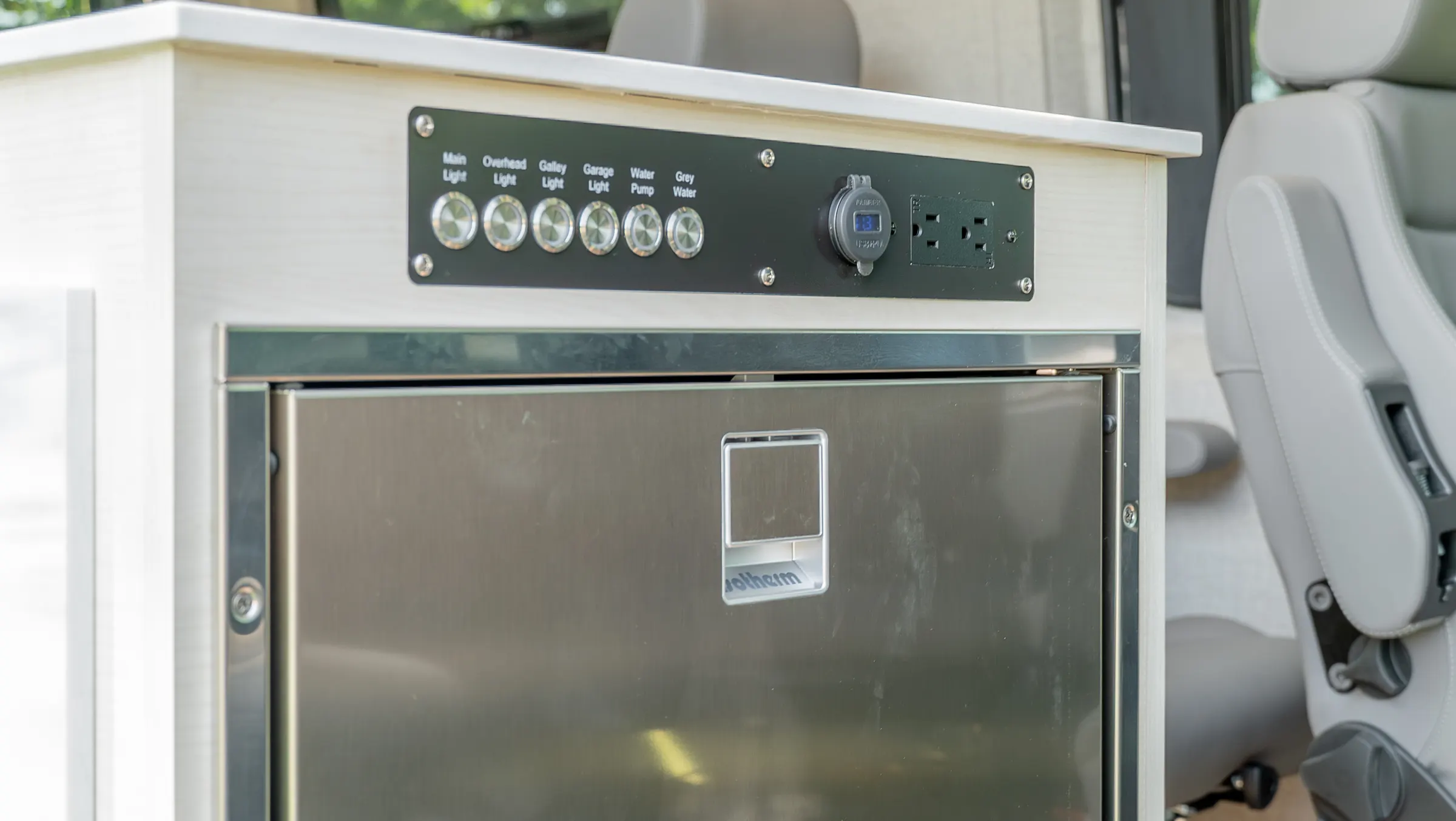 Stainless front-opening fridge in the galley cabinet, under a switch panel labelled for the lights, the water pump and the grey tank, with a twelve-volt socket and two outlets