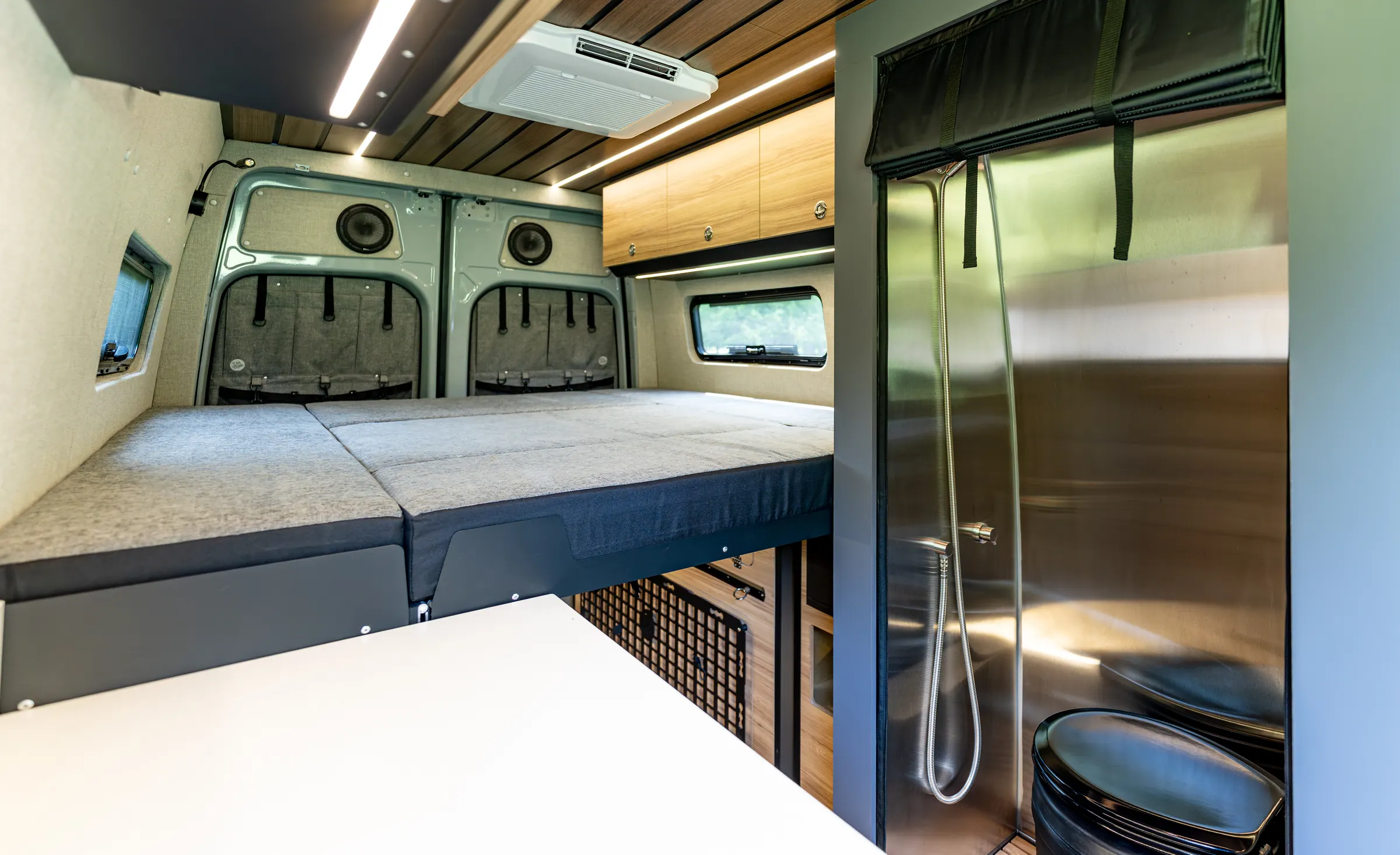 The rear bed made up beside the stainless-lined wet bath, the hand shower on its hose and the toilet below it, with the rear doors and their mesh gear panels behind