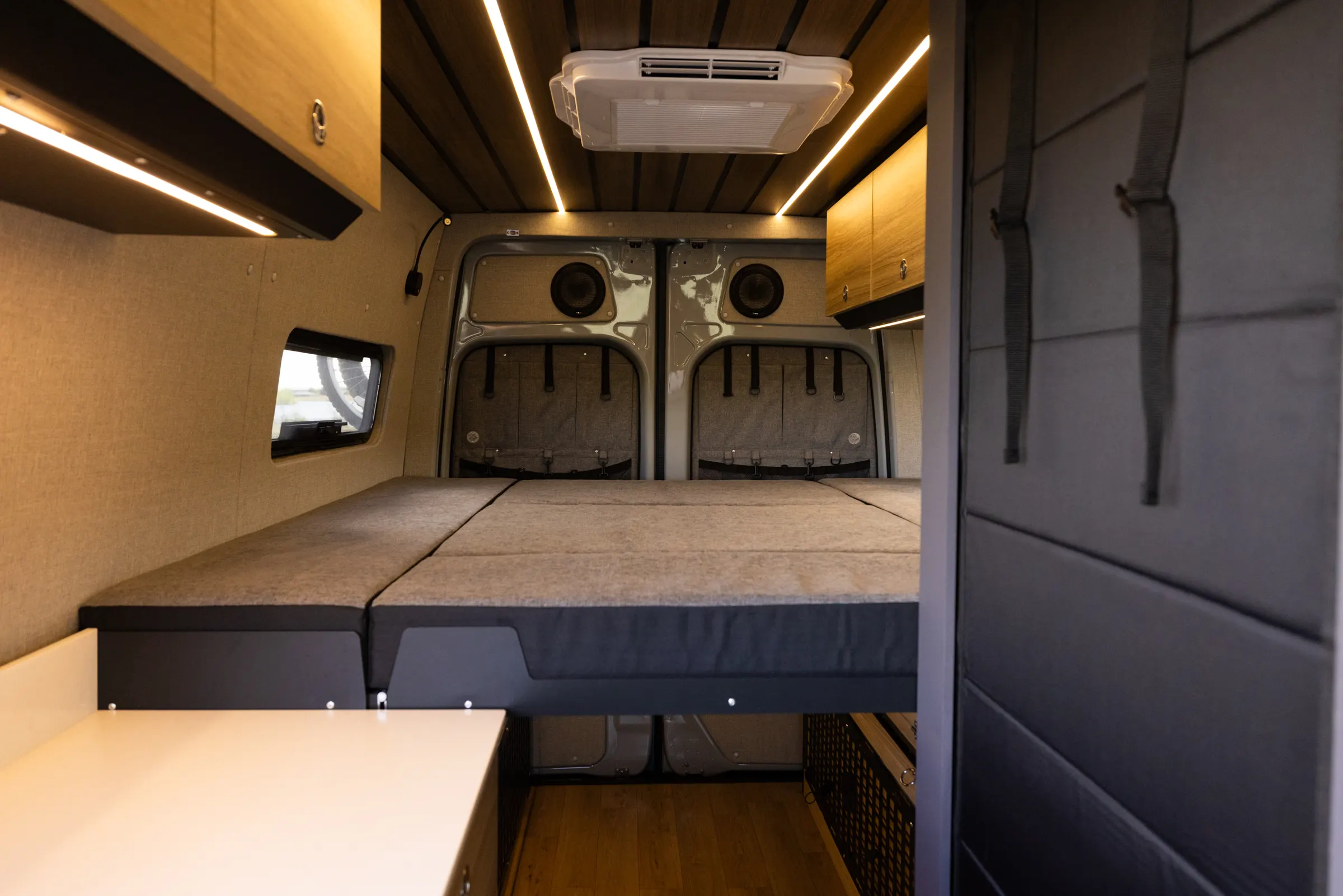 The rear bed made up across the full width of the cabin, the rear doors behind it carrying mesh gear panels, under overhead cabinets and an LED strip in the slatted ceiling
