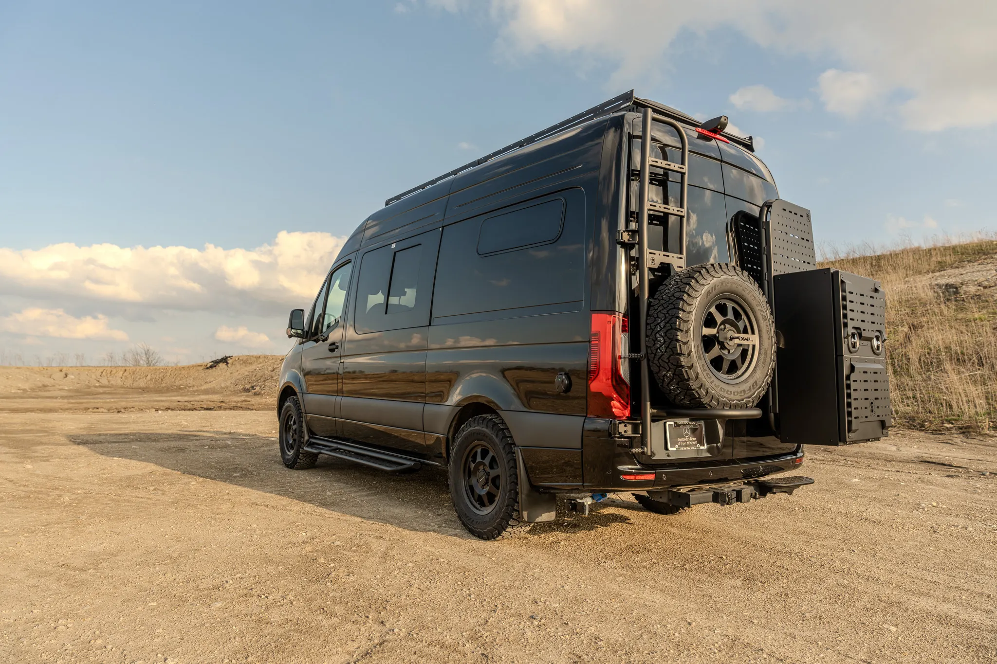 Rear three-quarter view on gravel, both rear carriers swung open — the spare wheel on one, the exterior cargo box on the other